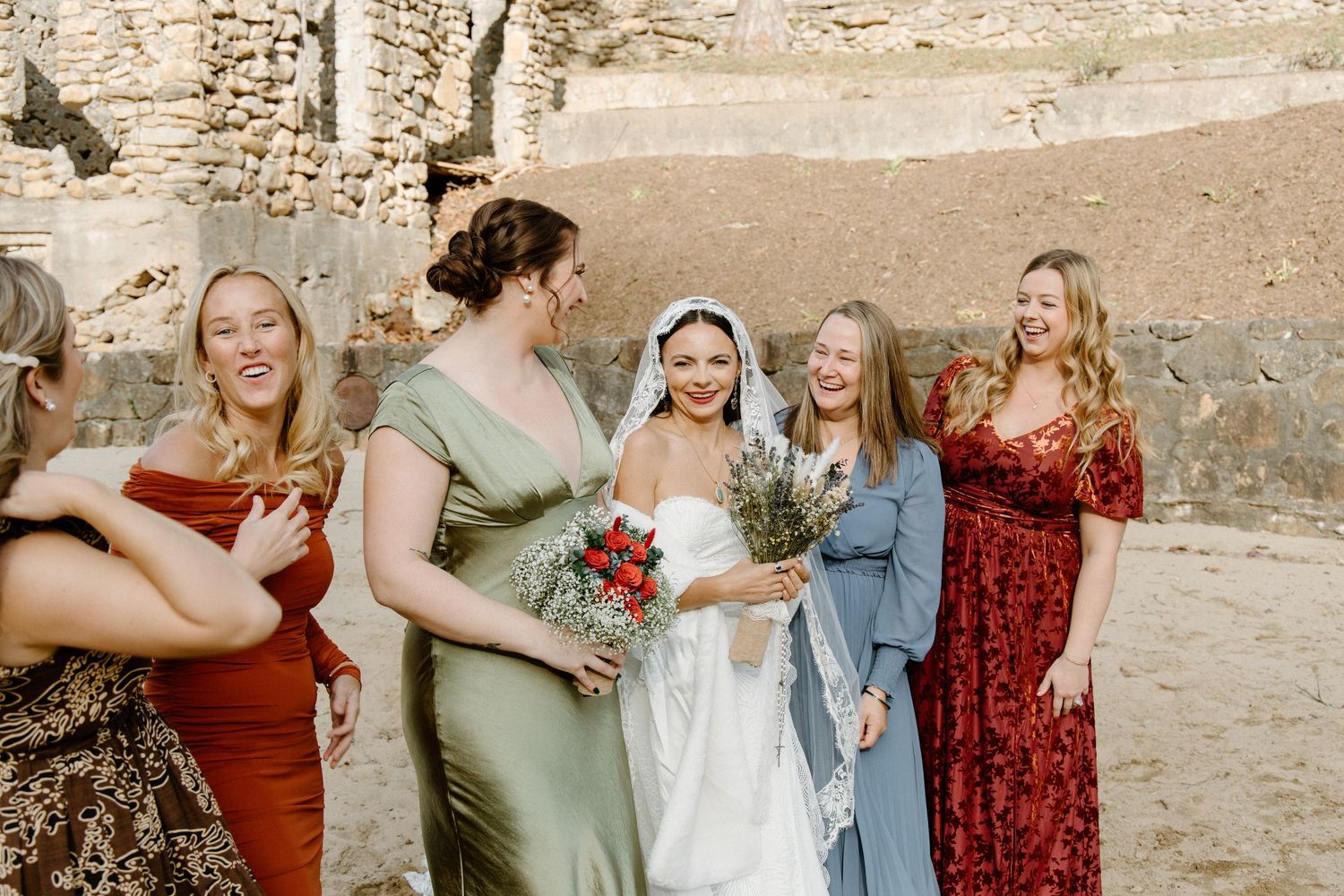 People in flowing dresses gather outdoors for a wedding celebration against a rustic stone wall background.