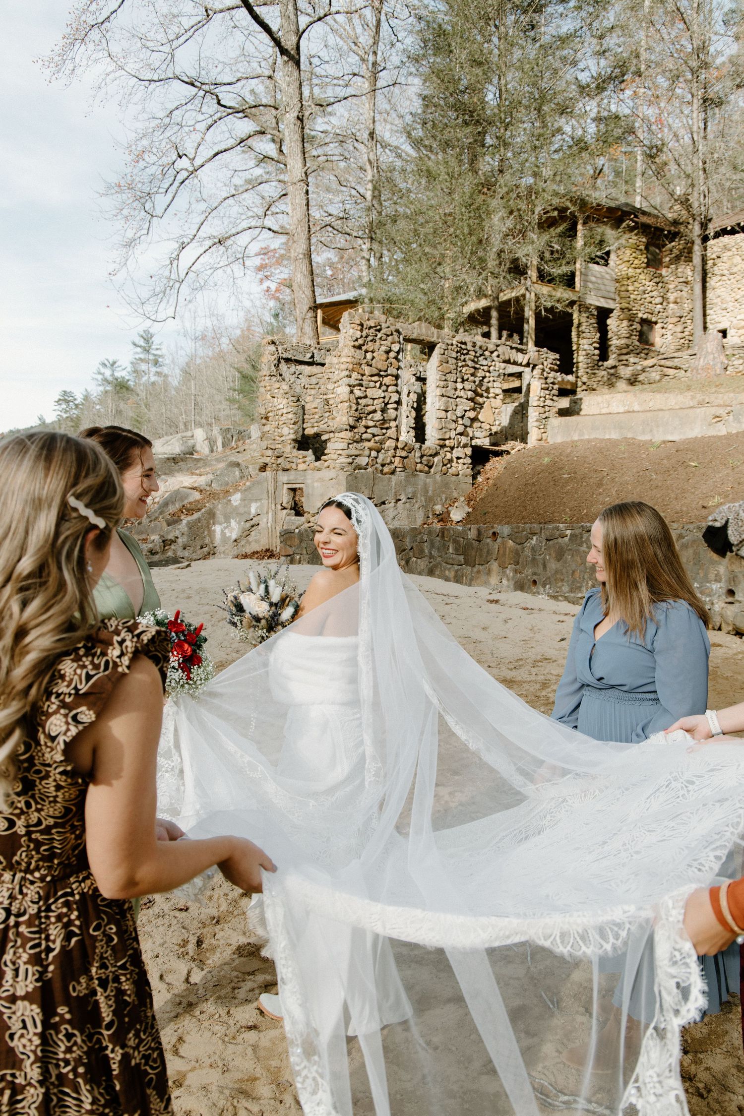 People outdoors help hold a flowing white wedding veil in the breeze near rustic stone buildings on a sunny day.
