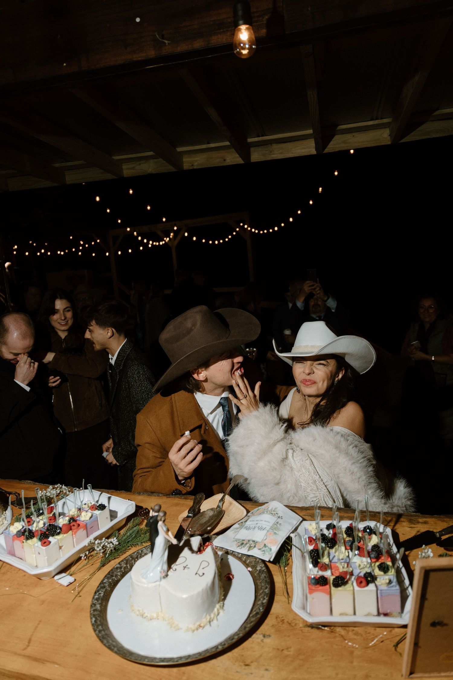 A western-themed wedding reception with cowboy hats and string lights shows a couple enjoying desserts at their table.