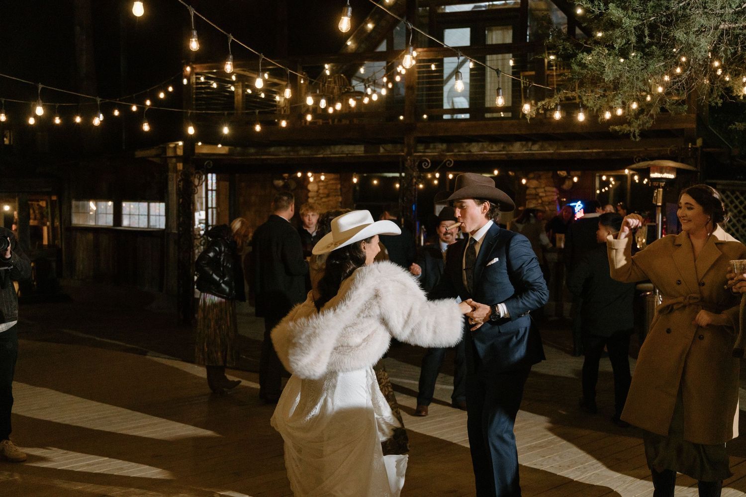 People dancing together at a rustic outdoor evening wedding reception under warm string lights.