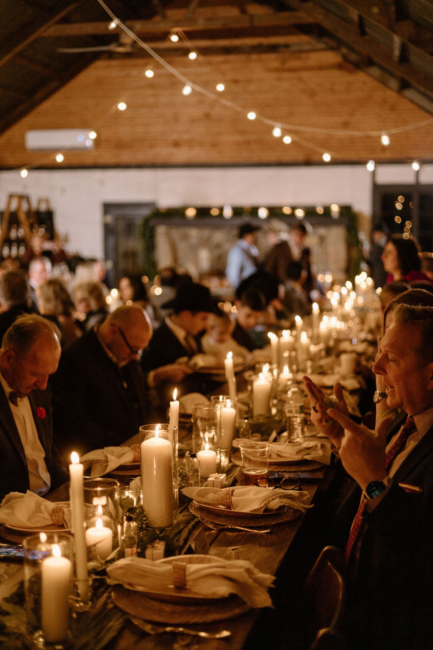 Guests enjoy an intimate candlelit dinner in a rustic barn venue with string lights overhead creating a warm ambiance.