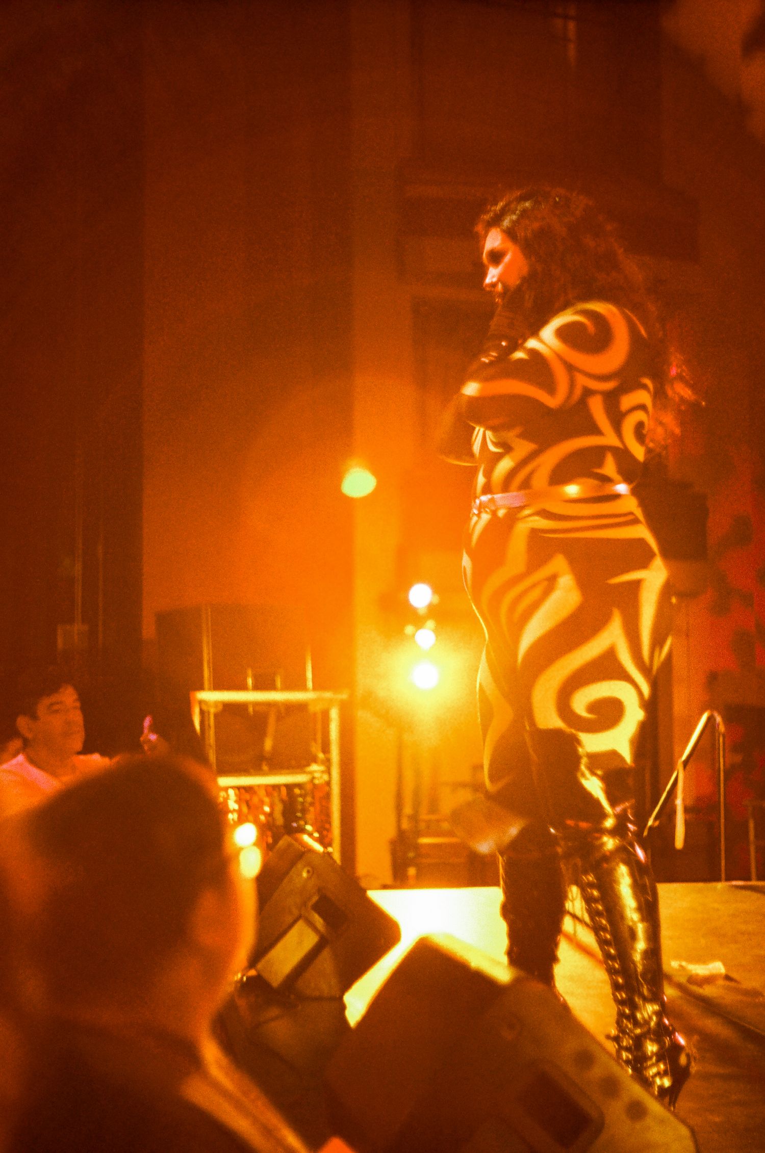 A performer in black and gold bodysuit stands on stage under warm orange lighting during a live performance.