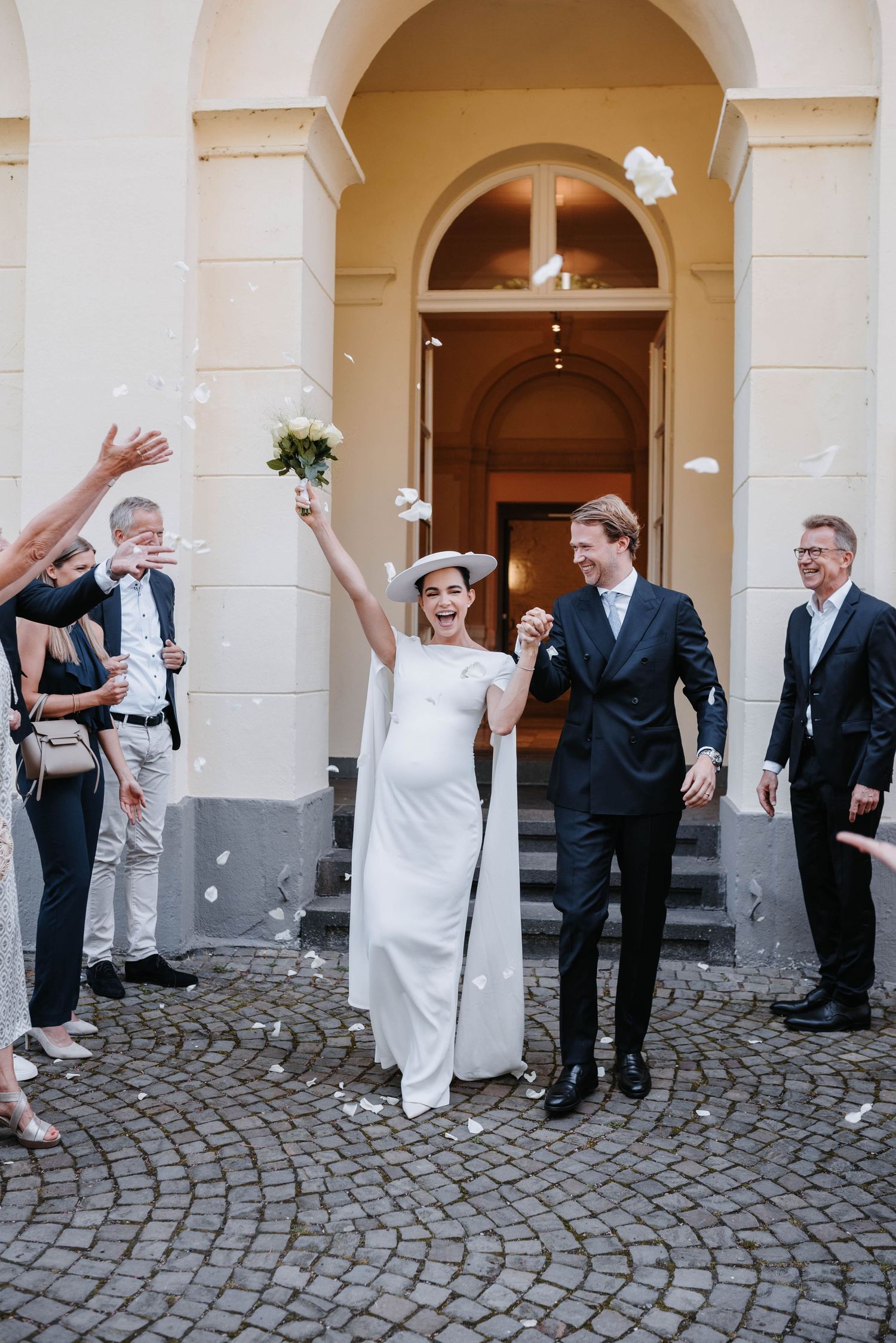 Wedding couple exits venue under archway while guests toss flower petals in celebration.