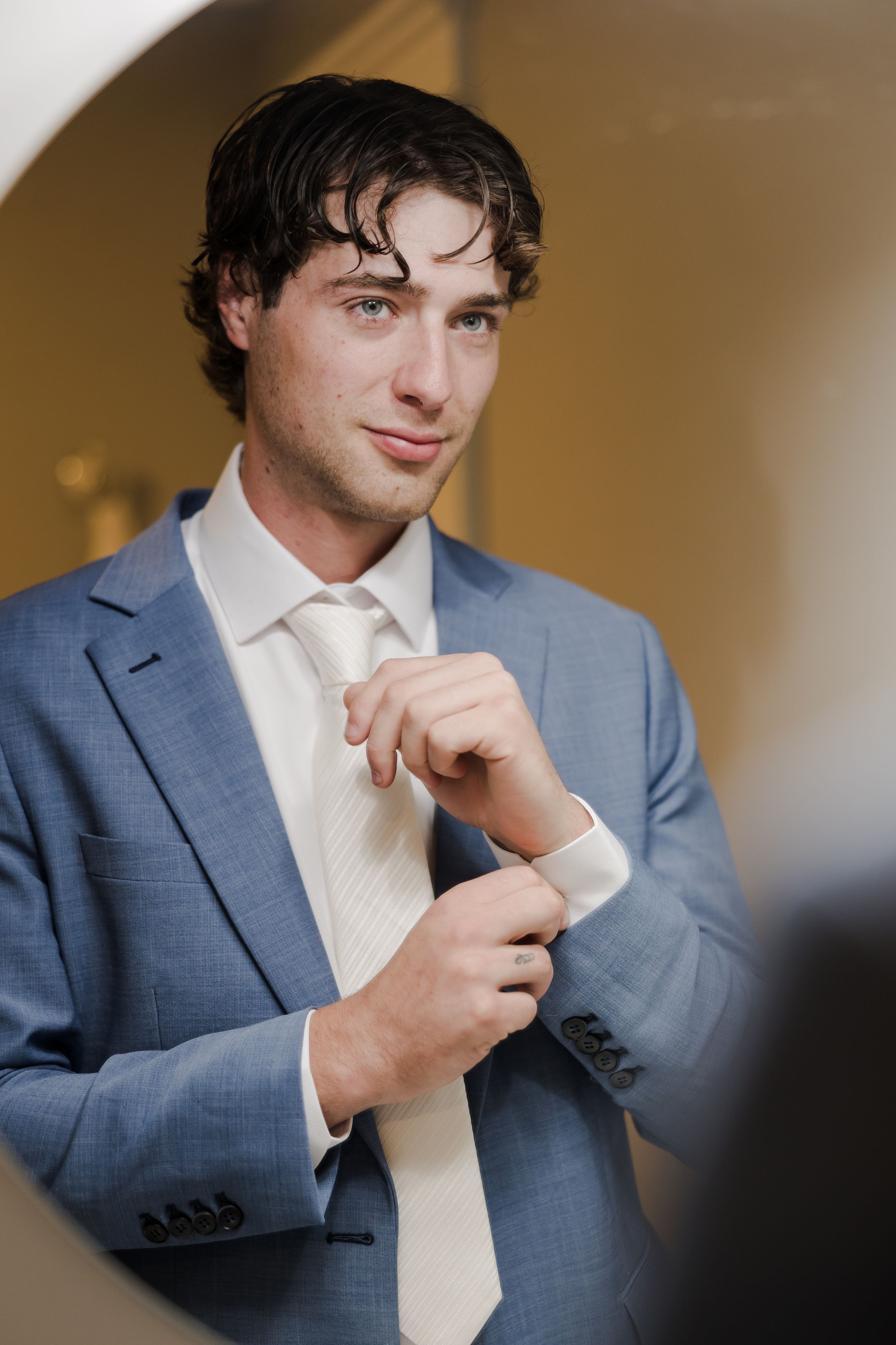 A person in a light blue suit adjusts their cream-colored tie while getting ready in a softly lit room.