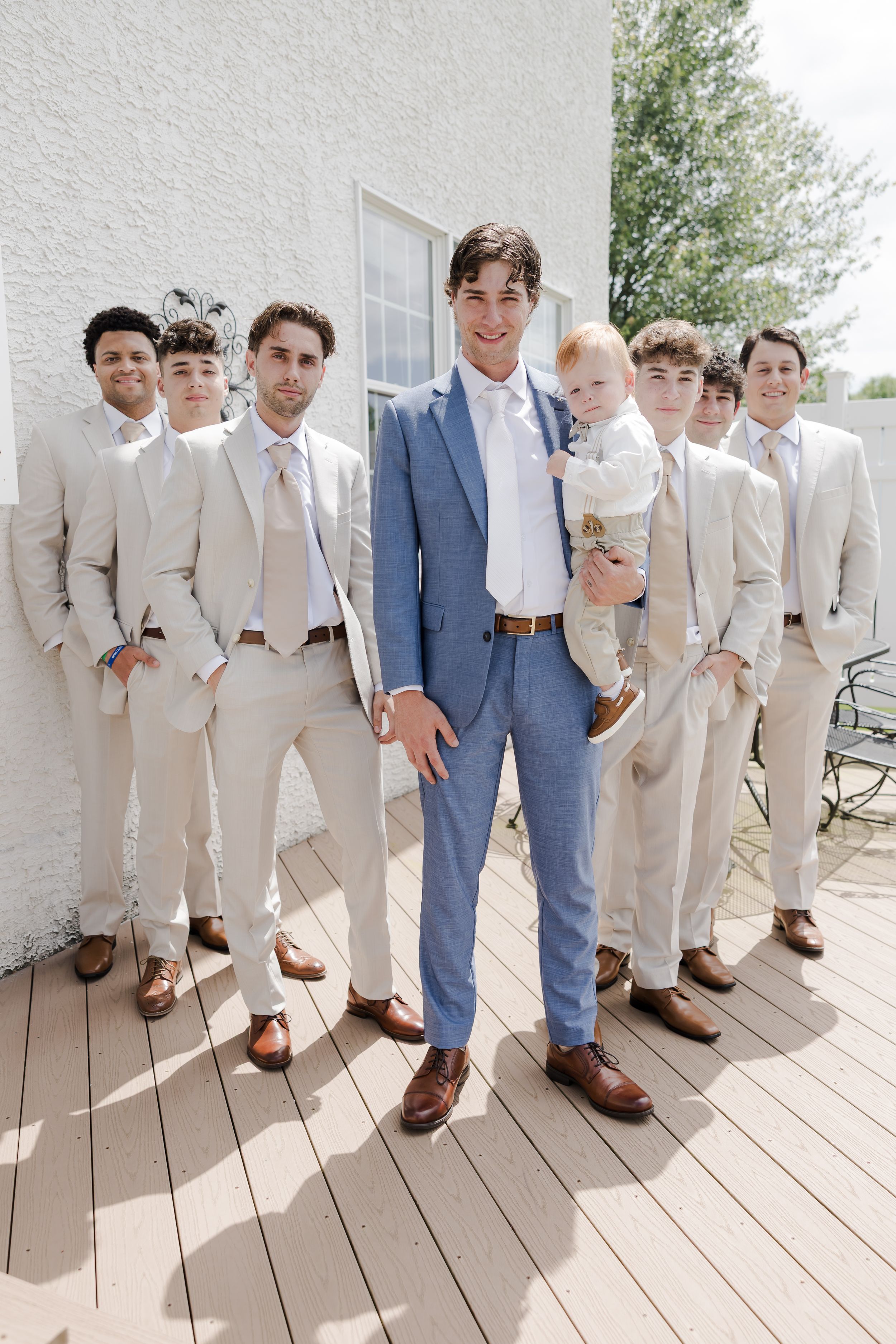 A groom in a blue suit stands with groomsmen in beige suits on a light wooden deck outside a white building.