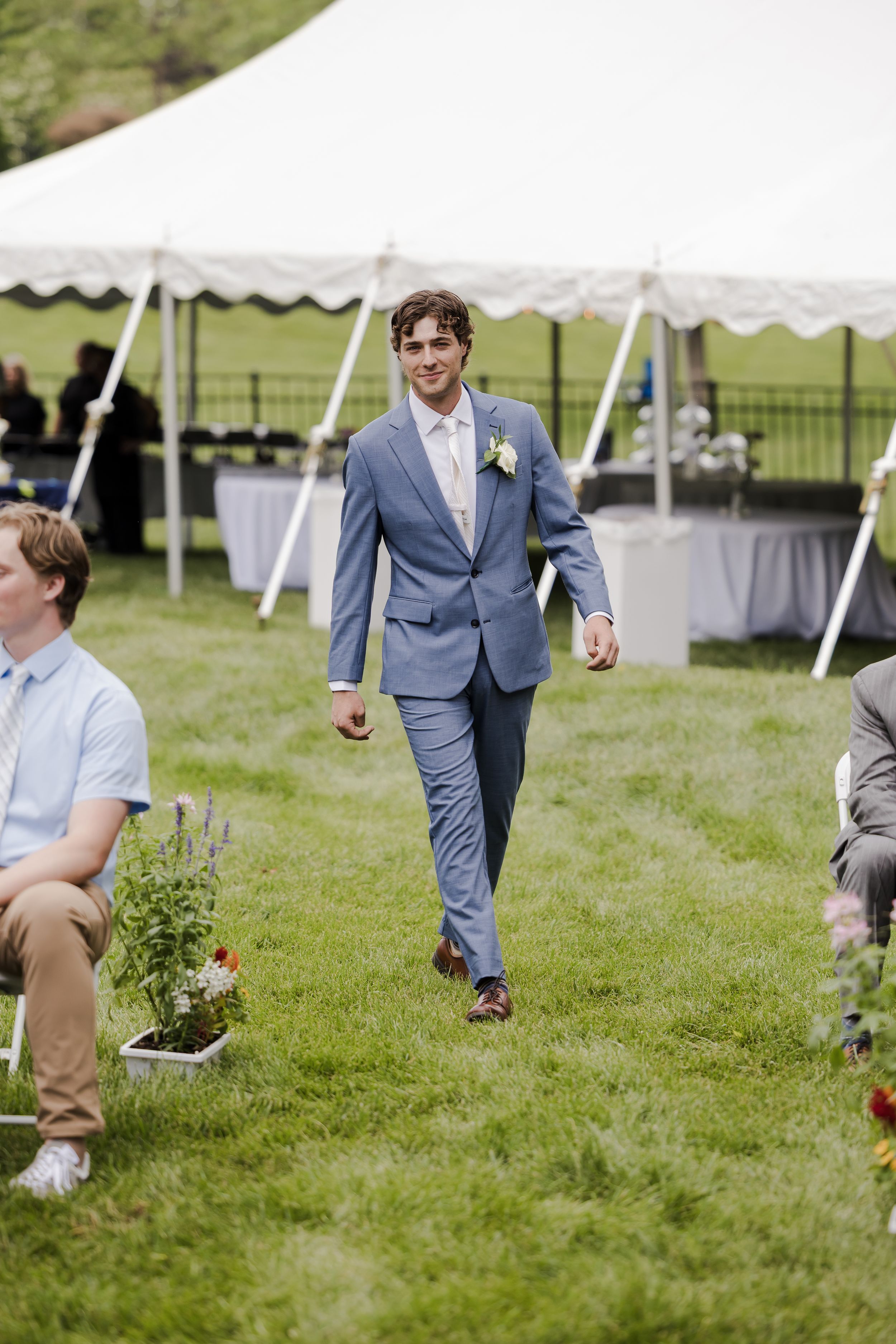 Wedding guest in a light blue suit walks across green grass with a white tent setup in the background.