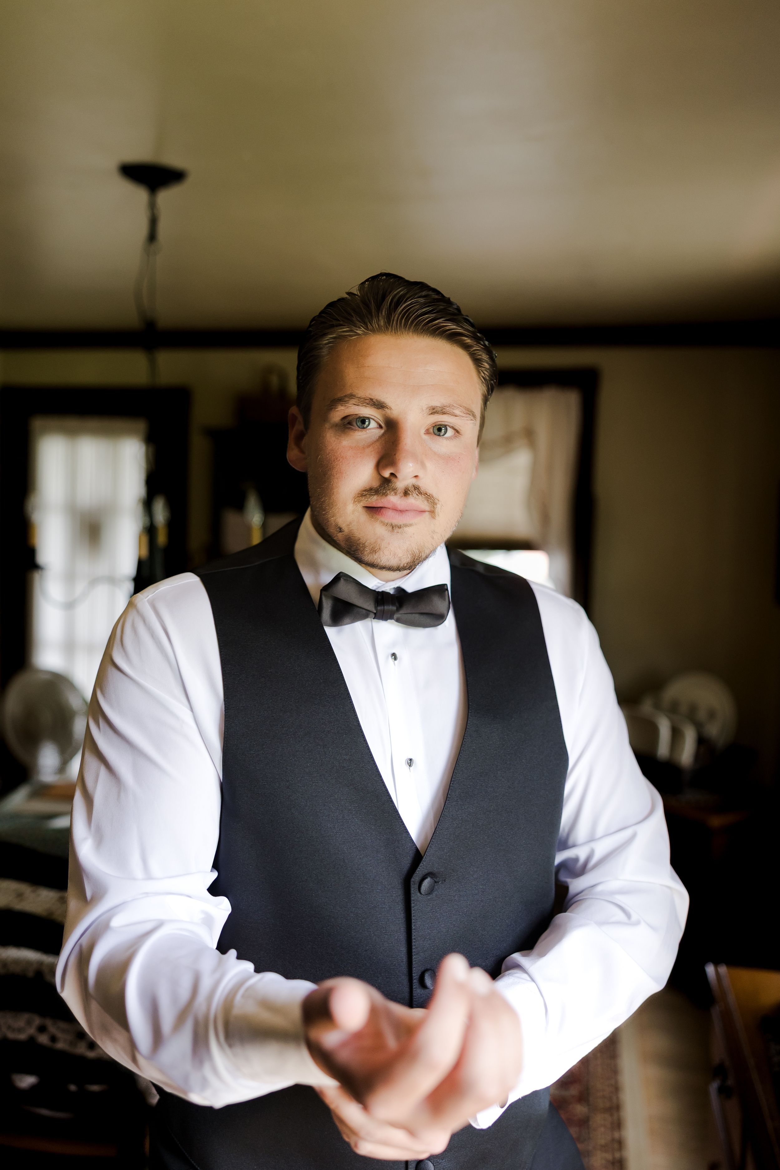 A person in formal attire adjusts their bowtie while wearing a black vest and white dress shirt in an interior setting.