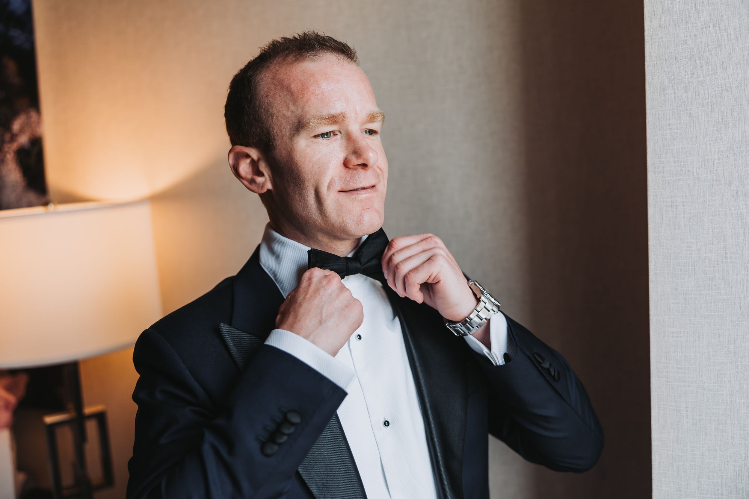 A person in a black tuxedo adjusts their bow tie while getting ready in a warmly lit room.