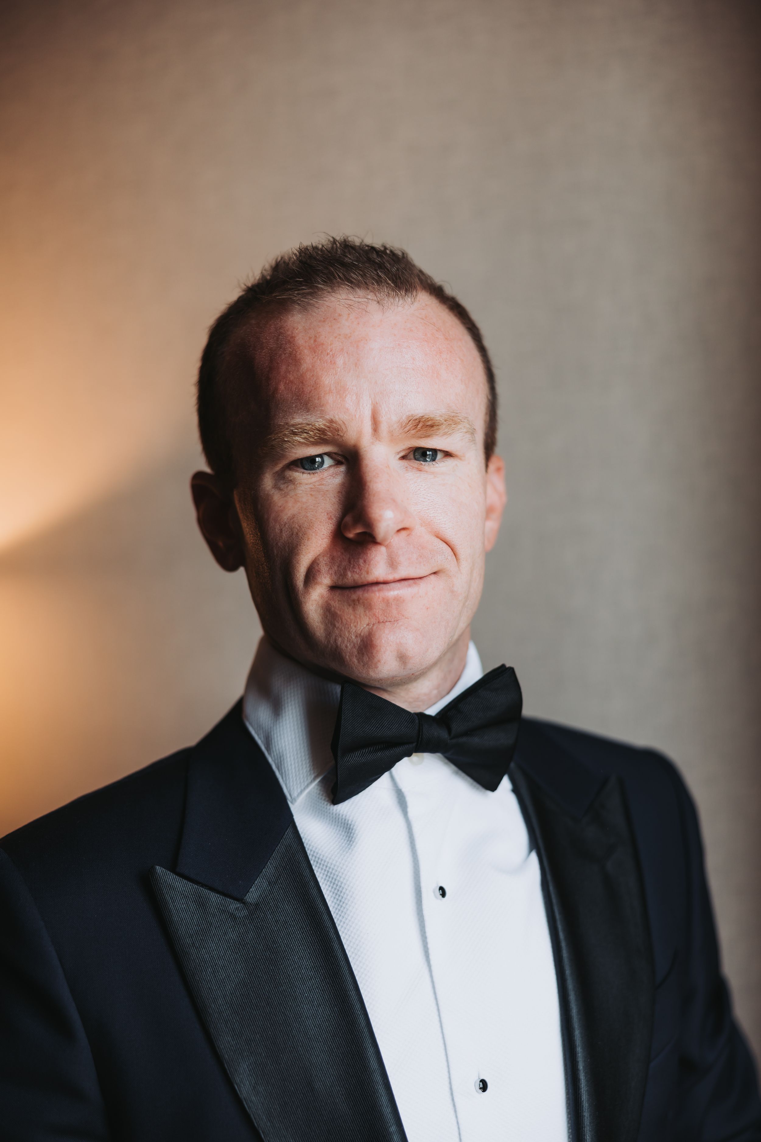 A person wearing a classic black tuxedo and bow tie smiles warmly in a formal portrait against a neutral background.