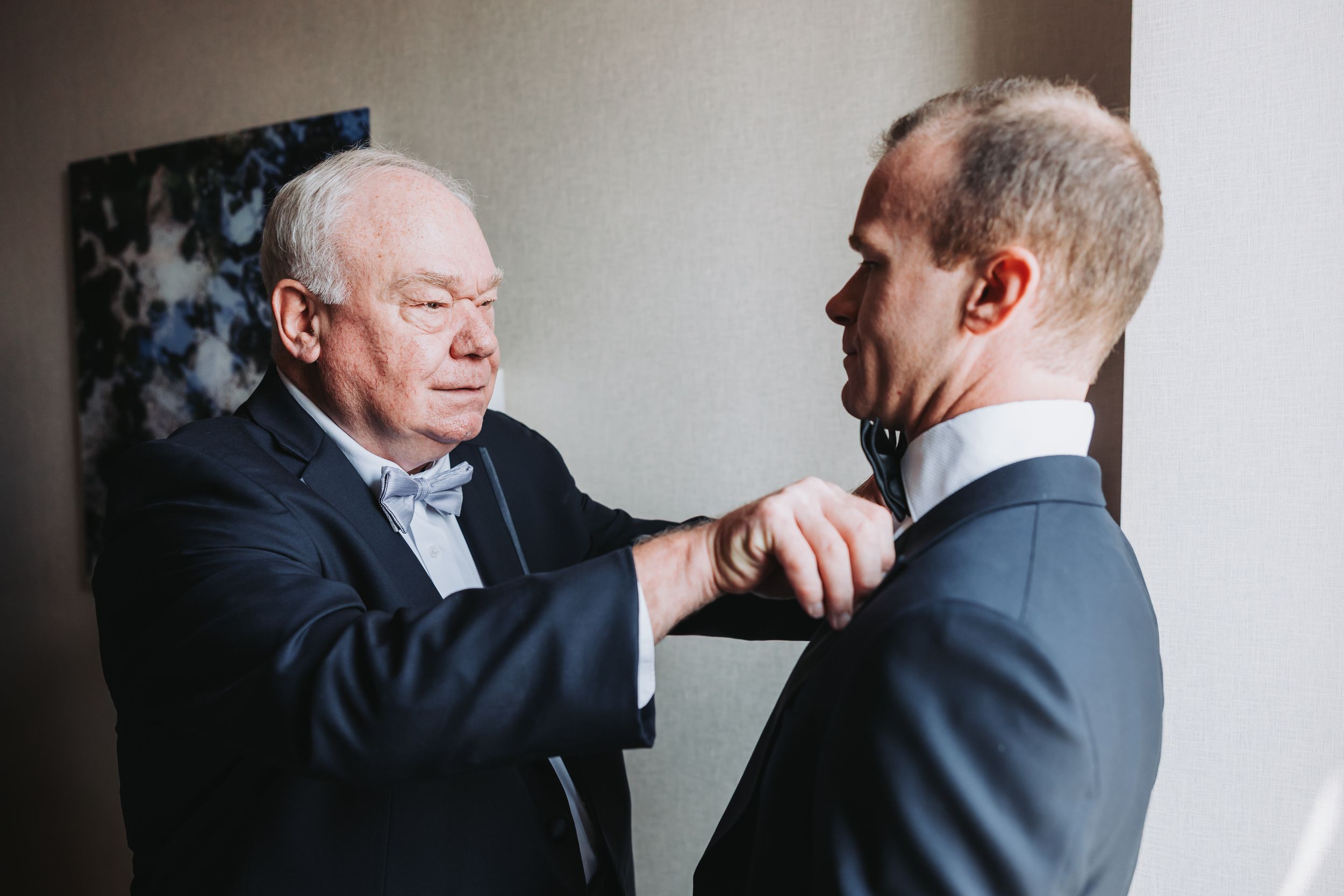 Two people in formal attire interact in an emotional moment against a white wall with artwork in the background.