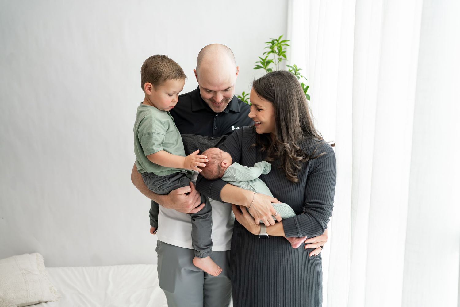 A family stands together in white room by window while looking down at their newborn baby.