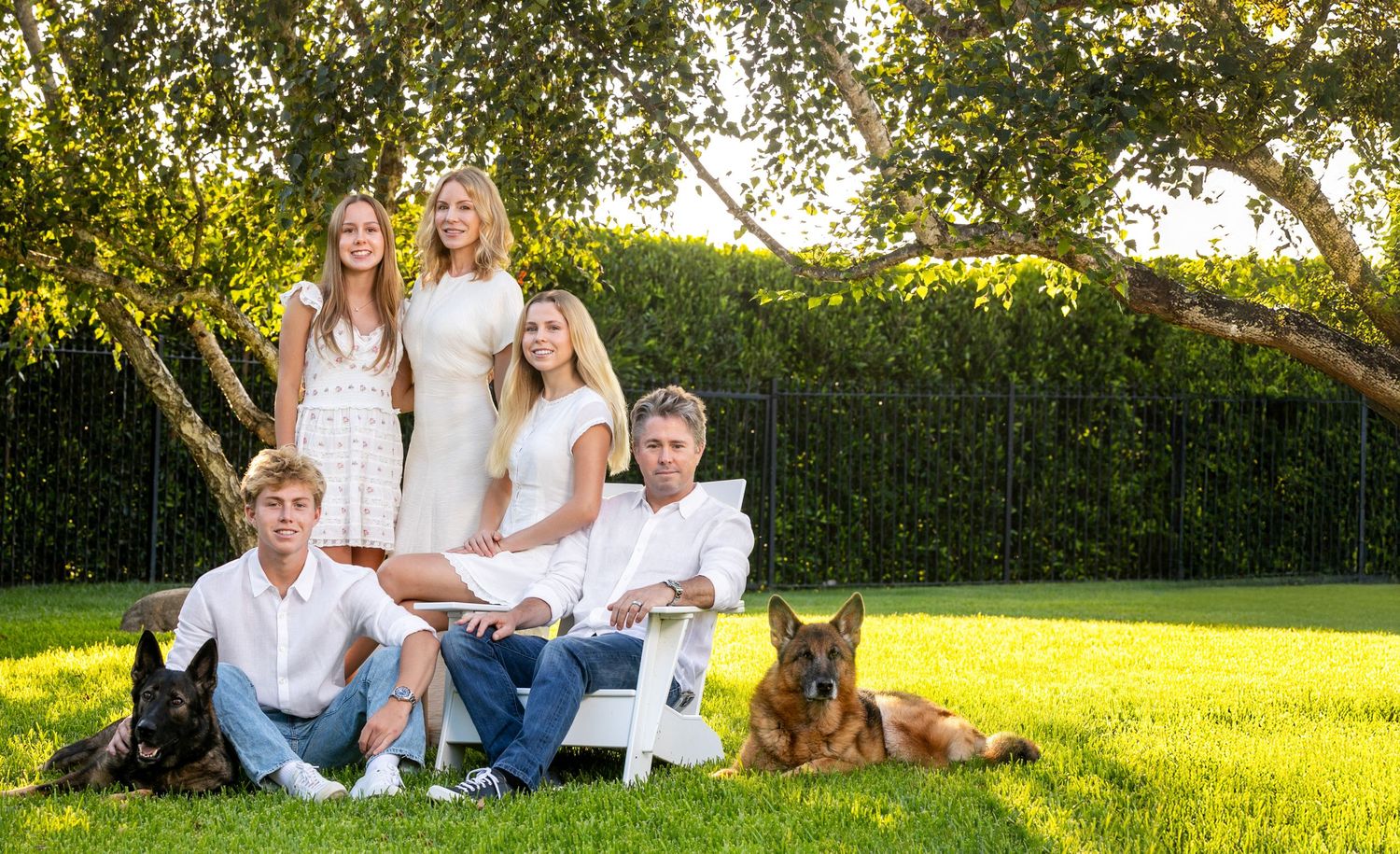 Family relaxing on white garden furniture outdoors with their two dogs on a sunny day with green lawn and hedges.
