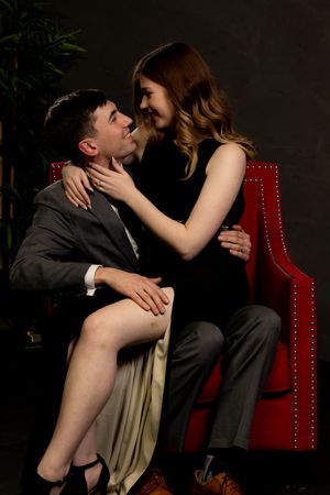 Intimate couple poses on red velvet chair in dark studio setting.