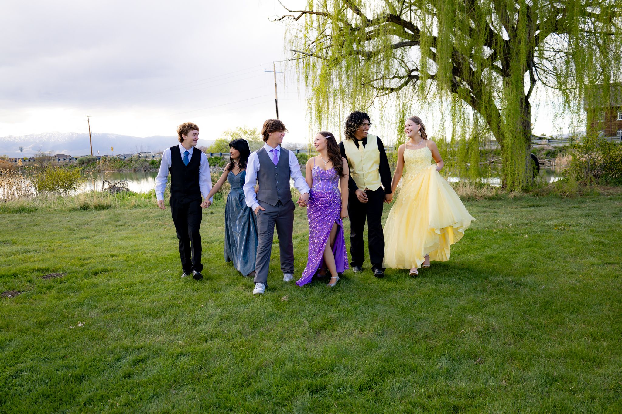 Formal group in colorful dresses and suits walking together on grass under willow tree.