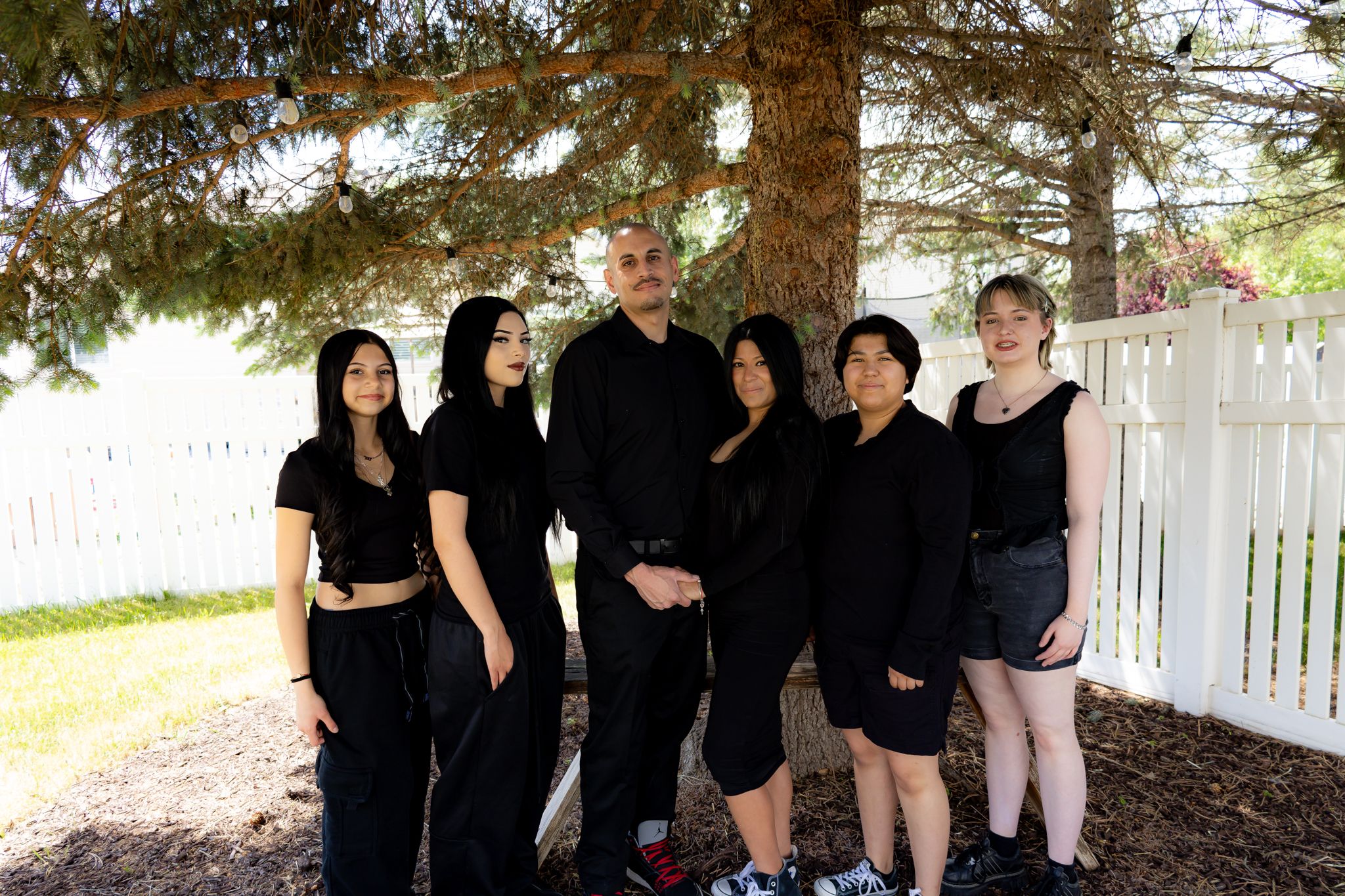 Group photo of people dressed in black outfits standing under a large pine tree.