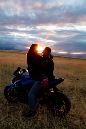 Silhouettes against setting sun with motorcycle in grassy field.