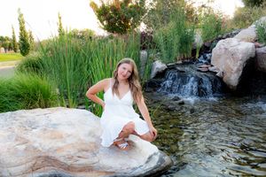 Woman in white dress sitting by waterfall and reeds during outdoor photoshoot.