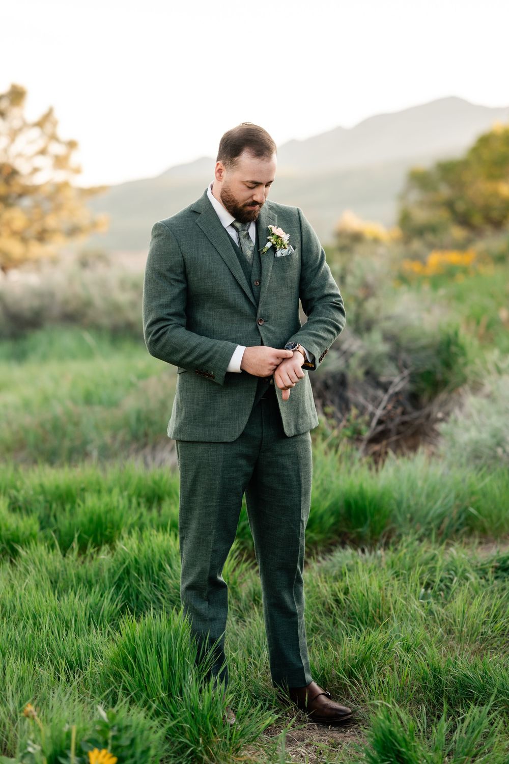 Groom in dark green suit checks his watch while standing in mountain meadow with yellow flowers.