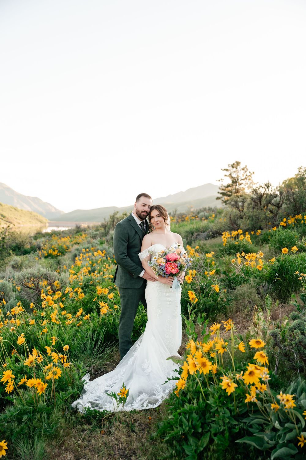 Wedding couple stands together in mountain wildflower field with lake view at sunset.