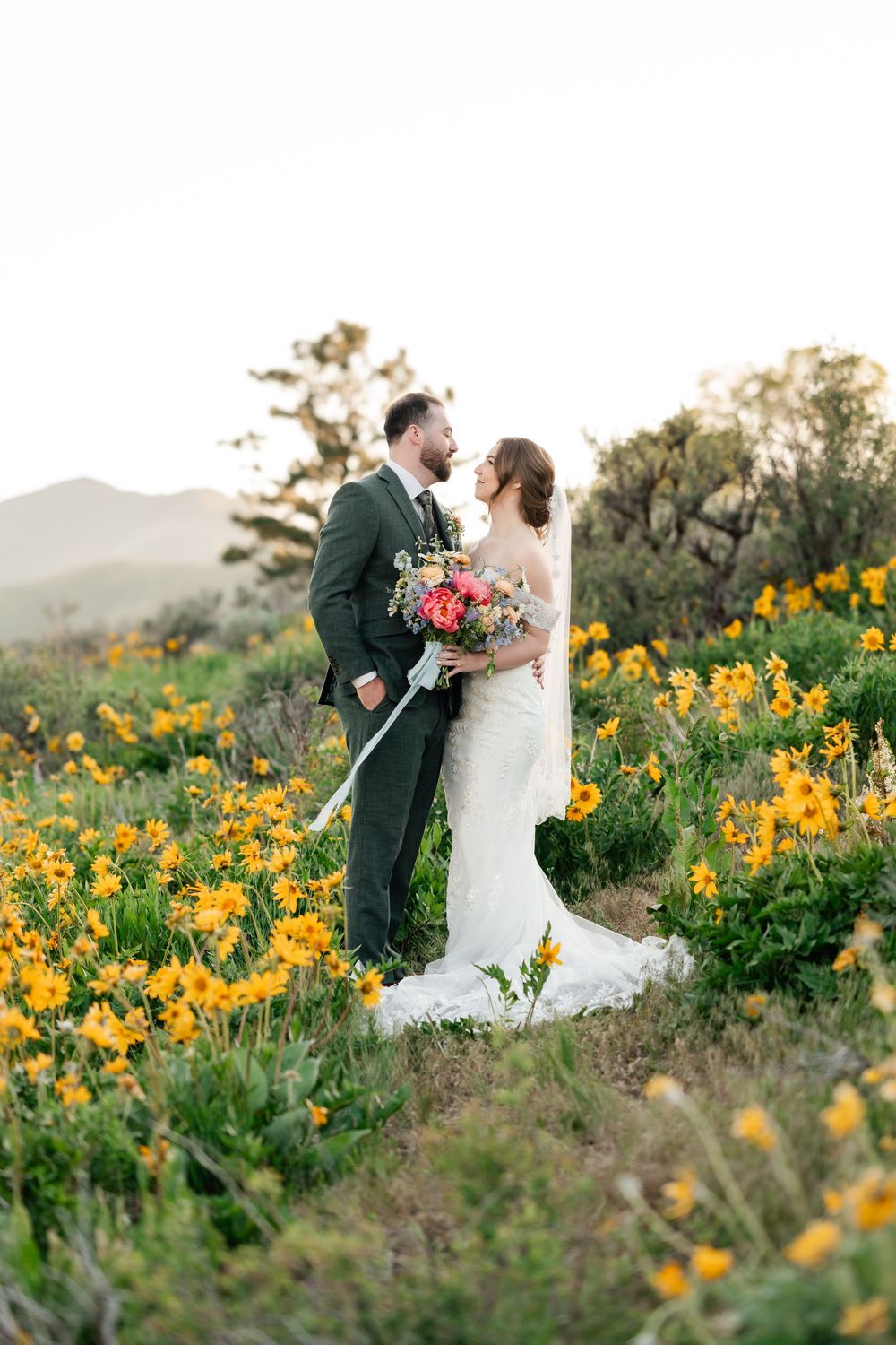 Romantic wedding couple embraces in a field of vibrant yellow wildflowers with mountains in background.