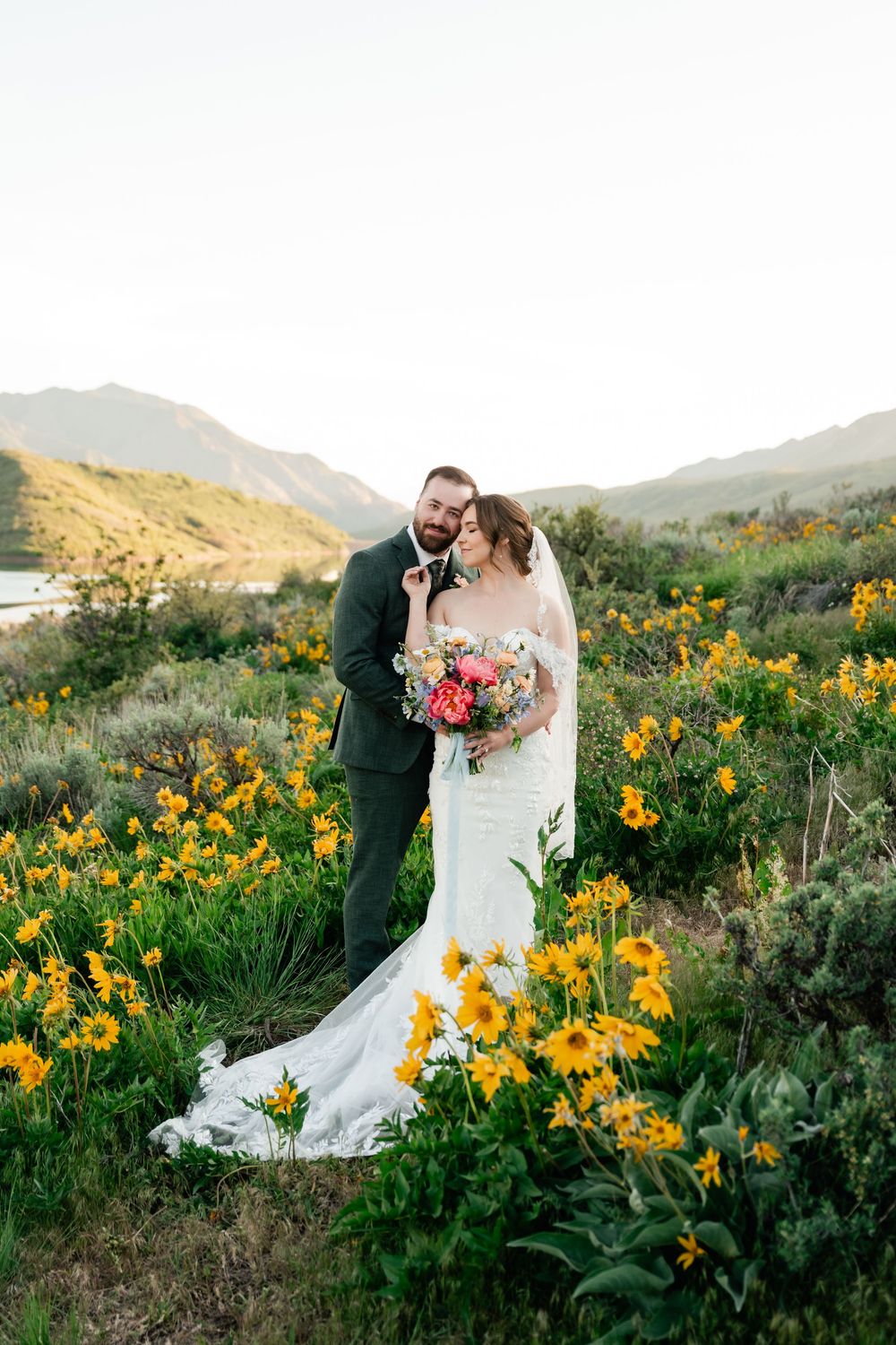 Newlyweds share intimate moment among yellow blooming flowers with mountain landscape behind them.