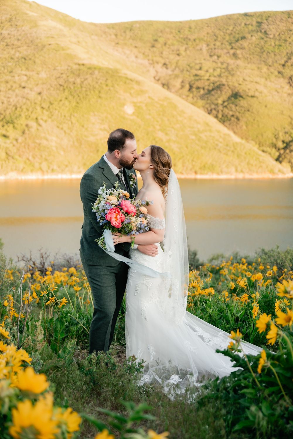 Bride and groom kiss beside lake with rolling hills and field of yellow flowers in beautiful natural setting.