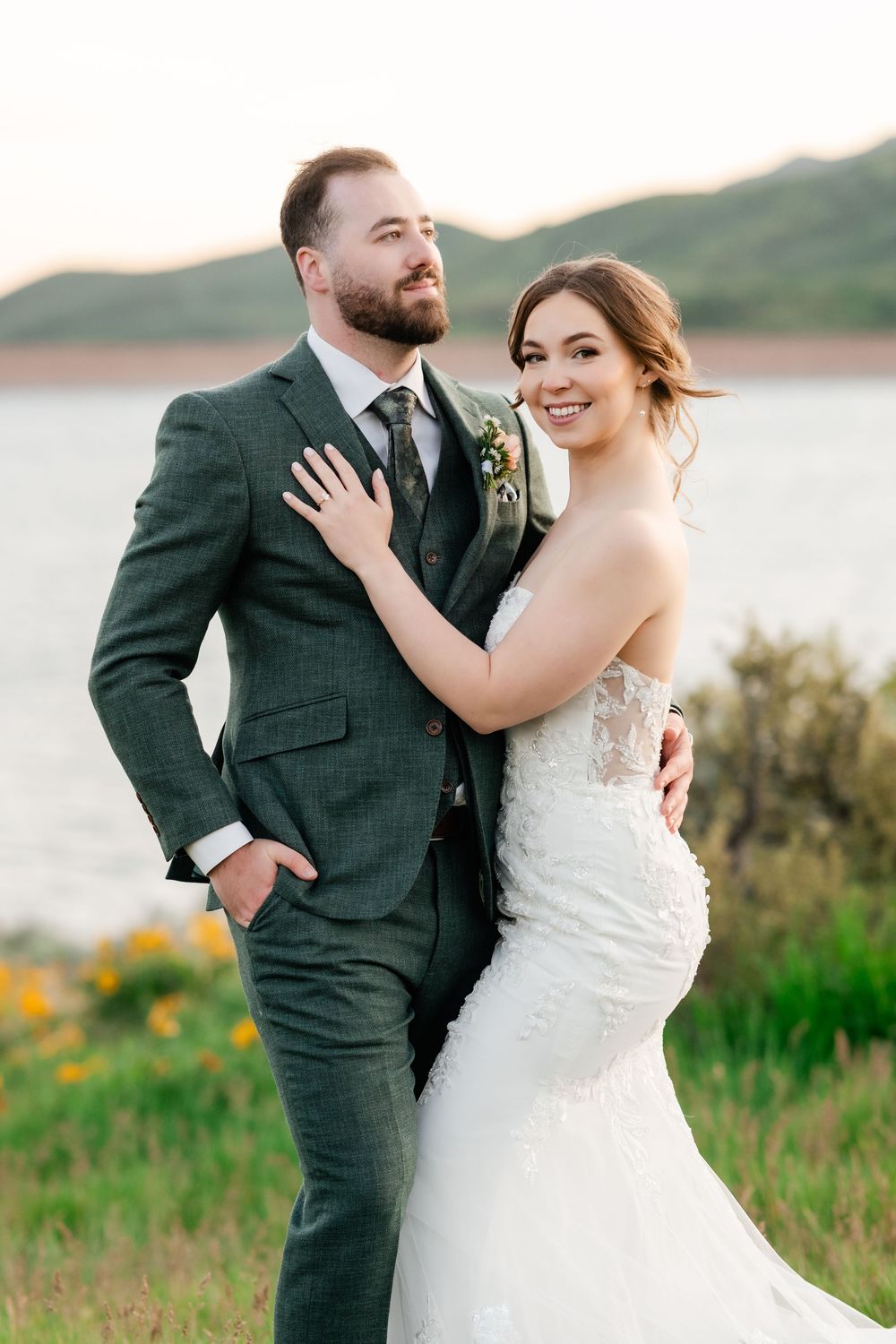 Wedding couple embracing by mountain lake with bride in lace dress and groom in dark green suit.