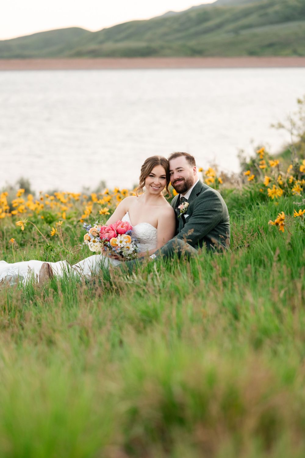 Intimate wedding portrait of couple lying in tall grass with lake and mountains in distance.