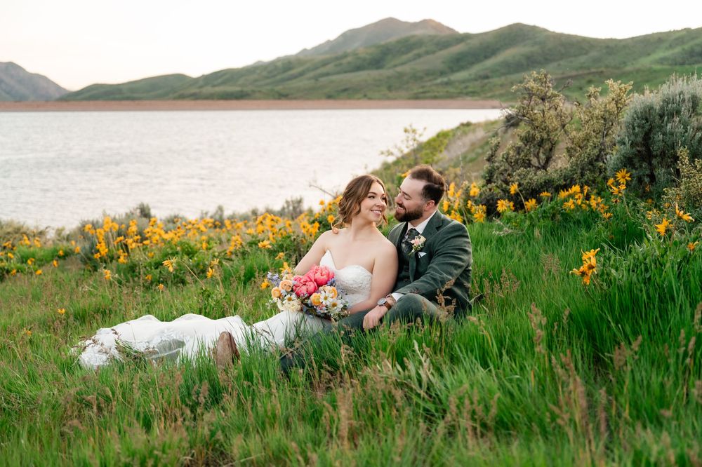 Couple sits in field of yellow wildflowers with mountain lake backdrop during sunset.