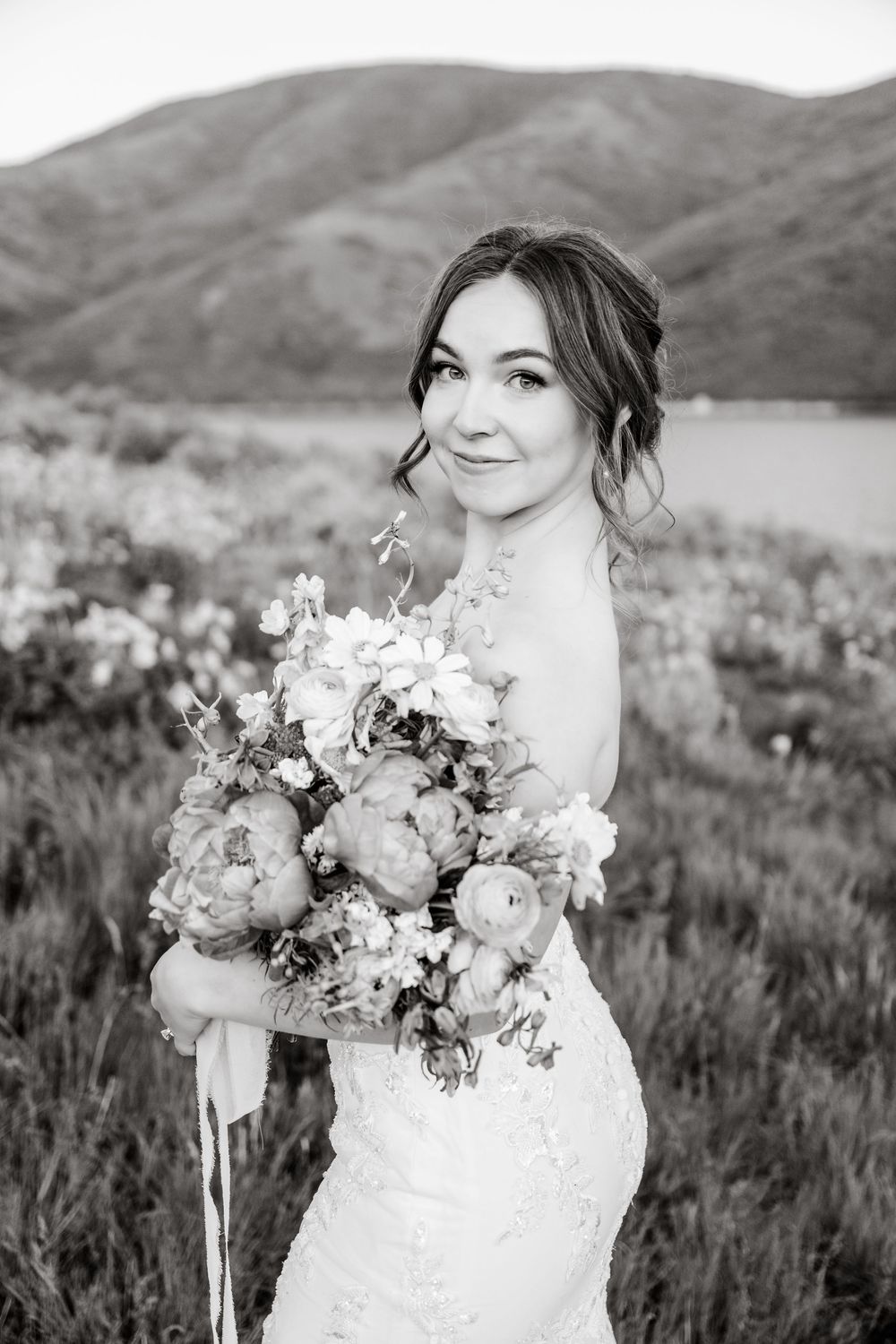 Bride in lace dress poses with elegant peony bouquet against mountainous landscape in black and white.