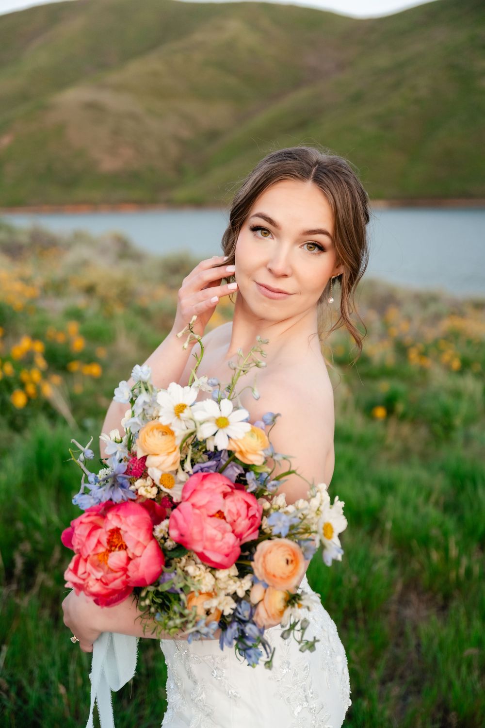 Bride holds colorful peony bouquet while posing in off-shoulder dress against mountain landscape.