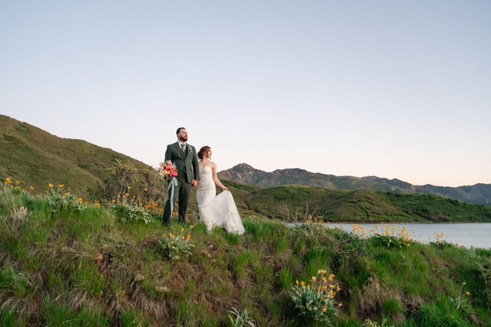 Wedding couple poses on hilltop at dusk with scenic mountain lake backdrop.