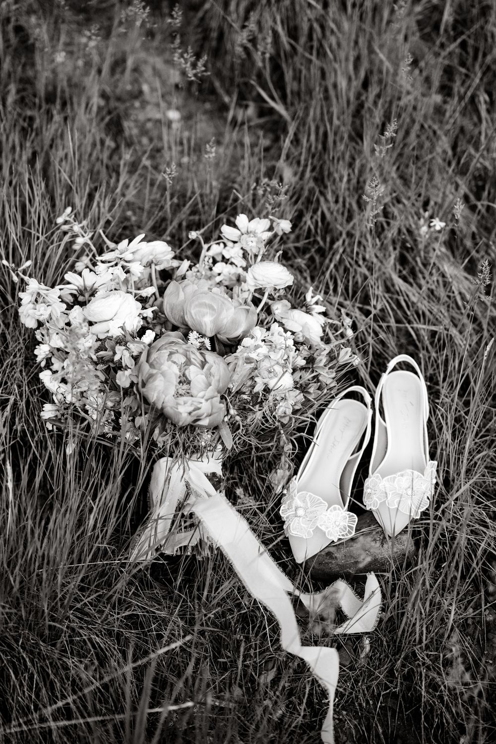 A black and white detail shot of elegant wedding shoes surrounded by delicate floral arrangements.