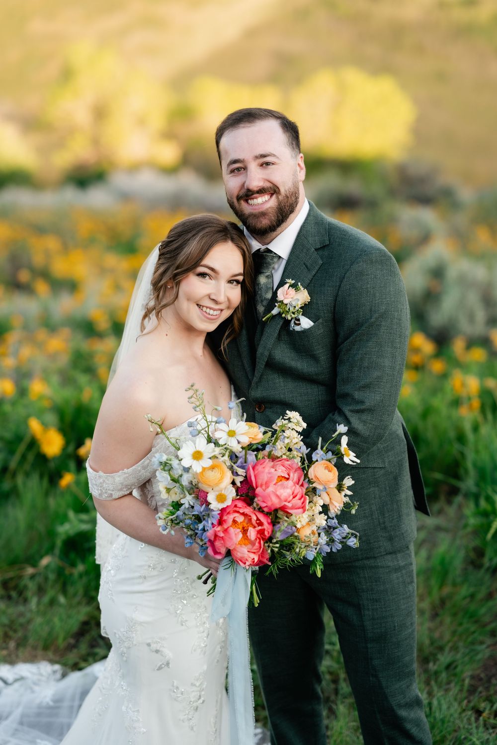Newlyweds pose in a field of wildflowers with vibrant coral and peach wedding bouquet against mountain backdrop.