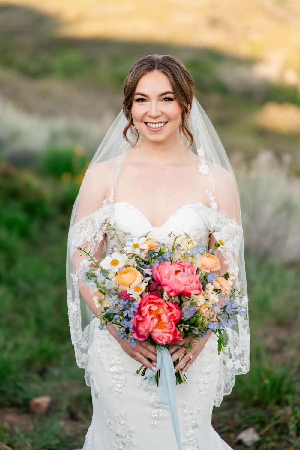 A bridal bouquet features vibrant coral peonies, white daisies, and delicate blue accents with flowing ribbons.