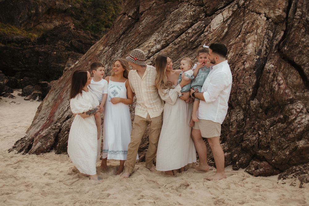 Group in white and beige attire gather for candid moment on sandy beach against dramatic rocky cliff backdrop.