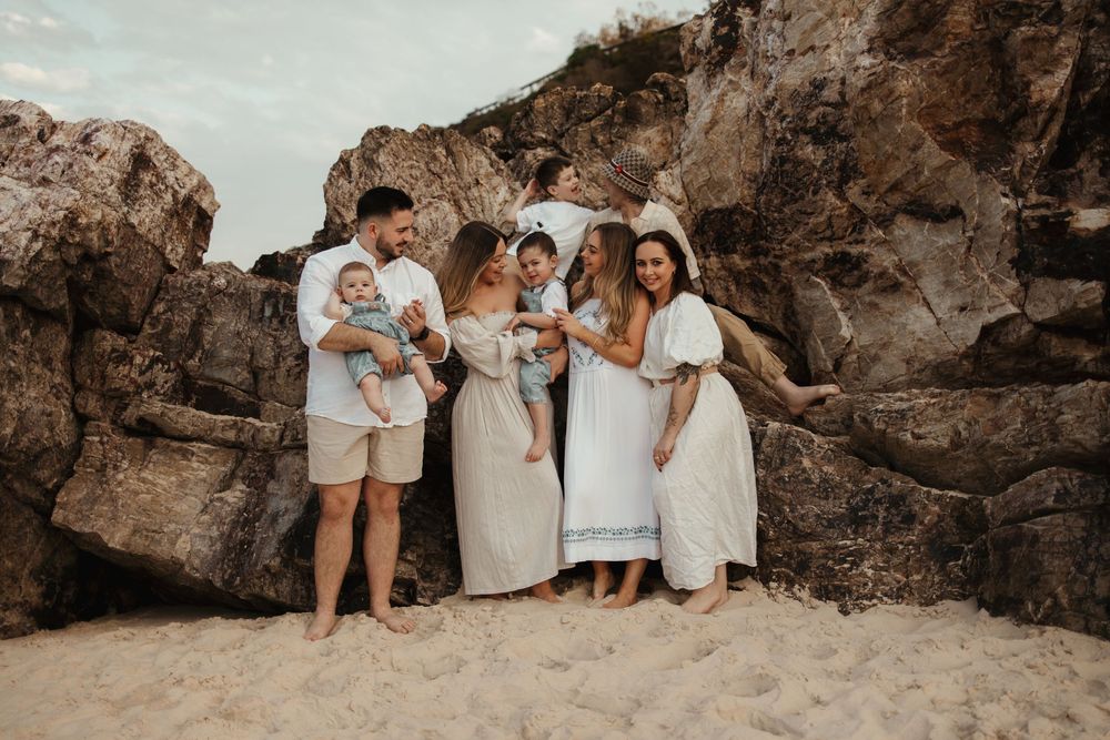 A group in coordinated white and neutral outfits poses together on a beach with dramatic rocky cliffs in the background.
