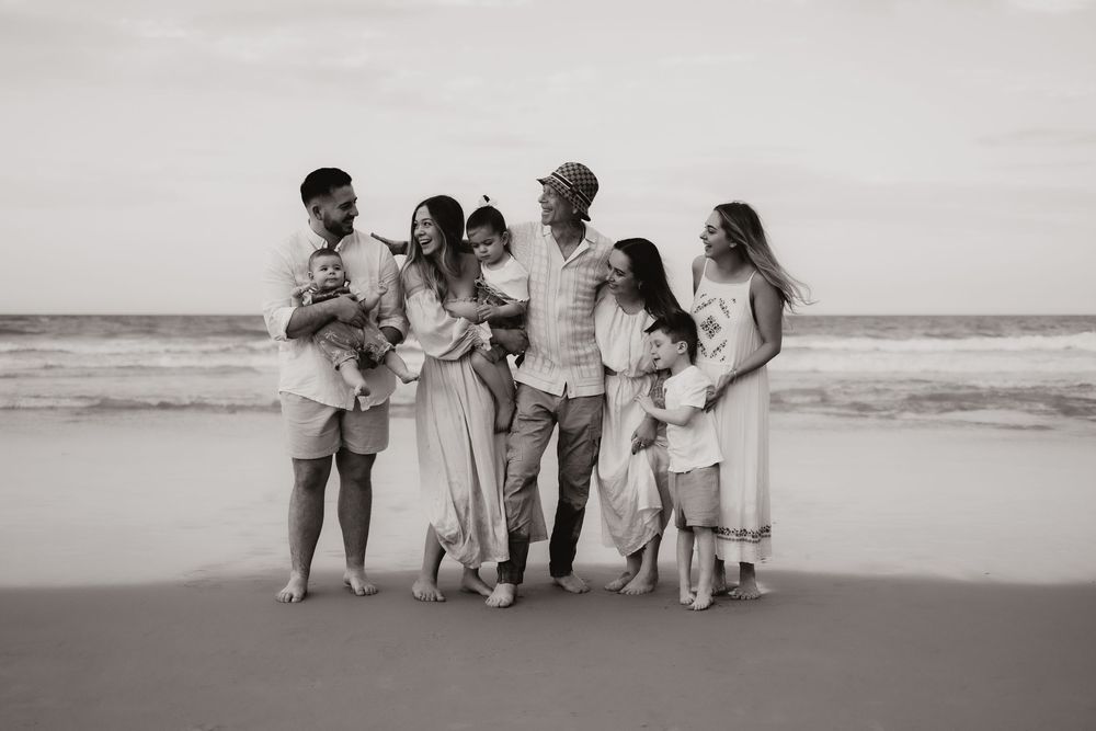 A group in coordinated light-colored outfits stand together on a beach shore in black and white photography.
