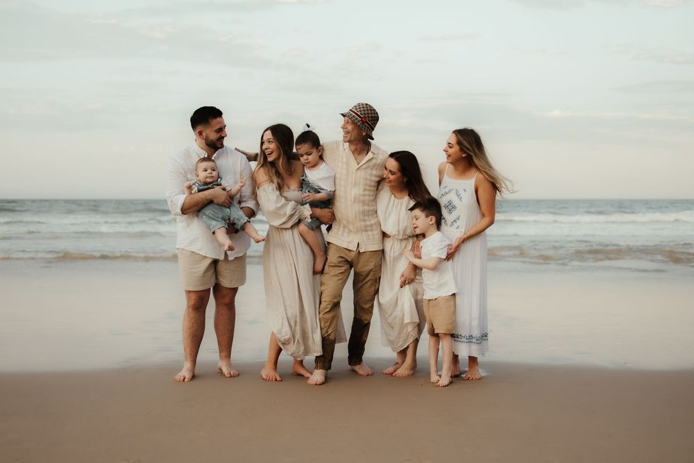 A family group in coordinated beige and white outfits stands together on a sandy beach at sunset.