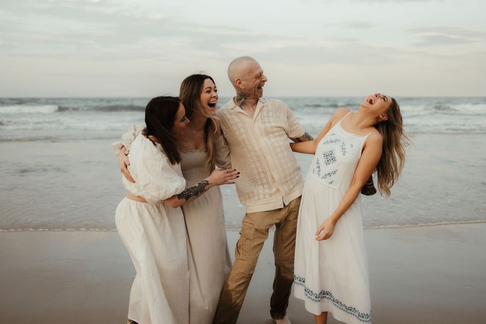 A group wearing white outfits laughs together joyfully while standing on a sandy beach at sunset.