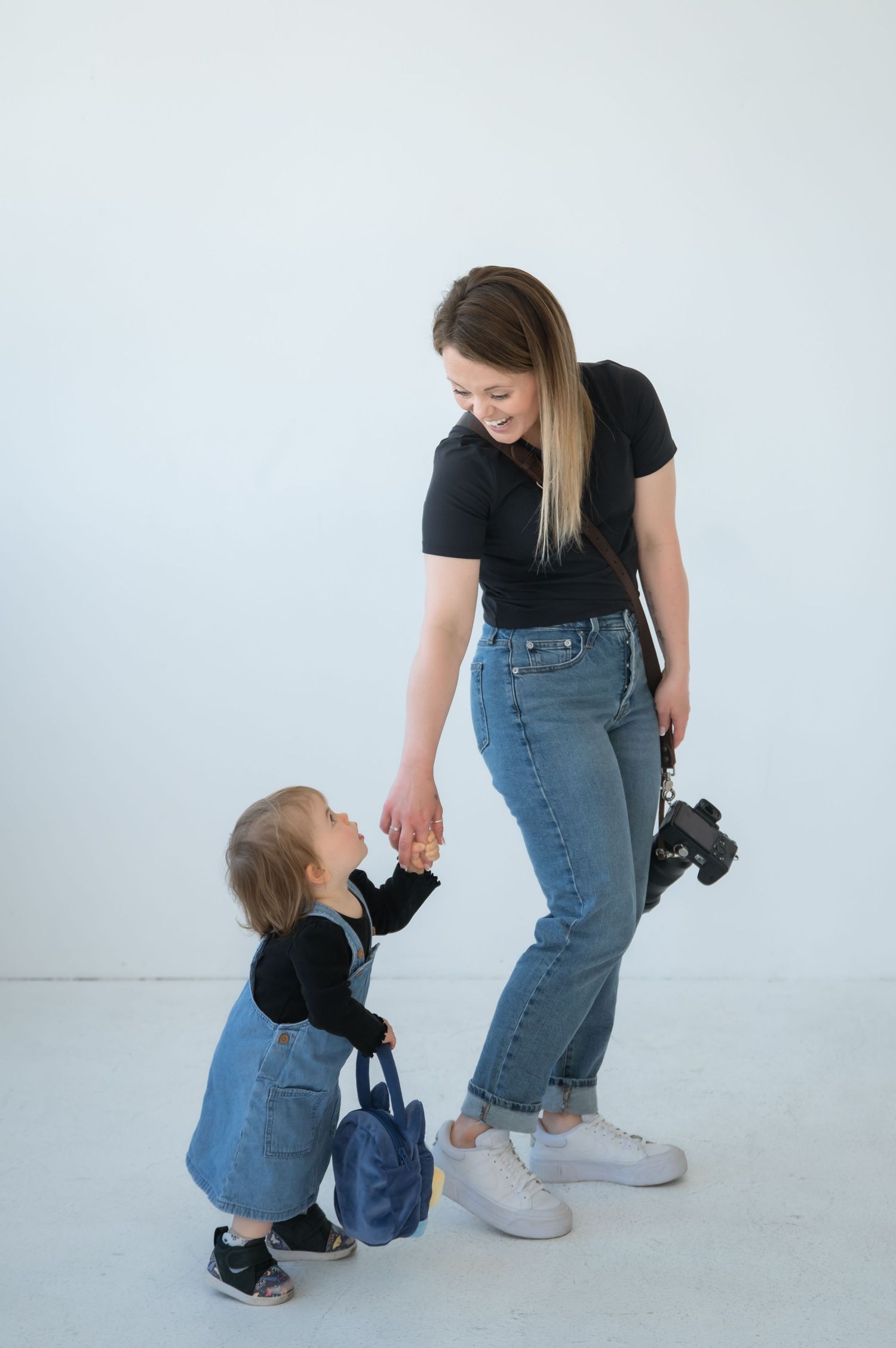 Woman in black shirt and jeans holding camera stands with matching denim-clad toddler in minimalist white studio setting.