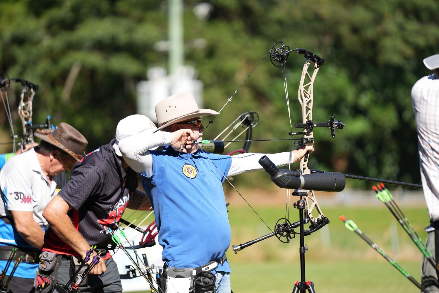 Archers in protective gear line up to shoot their compound bows at a competition on a sunny day.