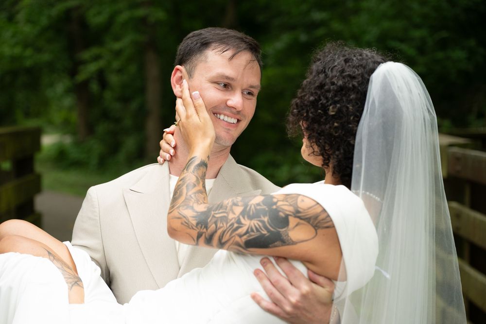 Groom with tattoos carries bride in white dress and veil, smiling outdoors at wedding.