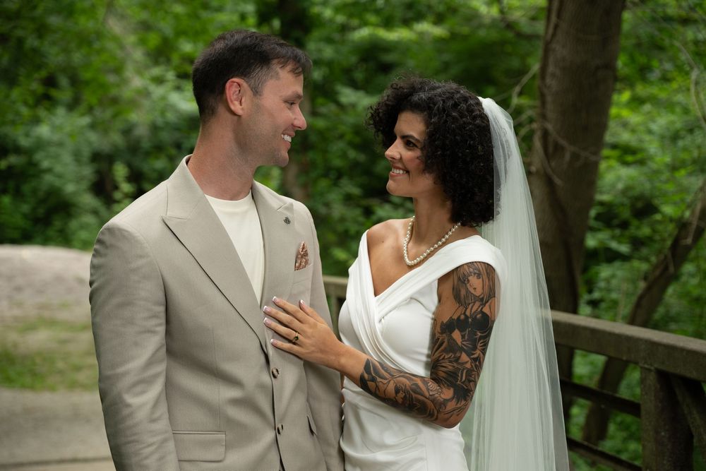Bride in white dress with sleeve tattoo and veil smiles at groom in tan suit outdoors in lush green park.