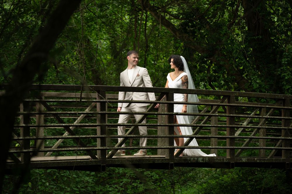 Bride and groom walking hand in hand on a wooden bridge surrounded by lush green trees.