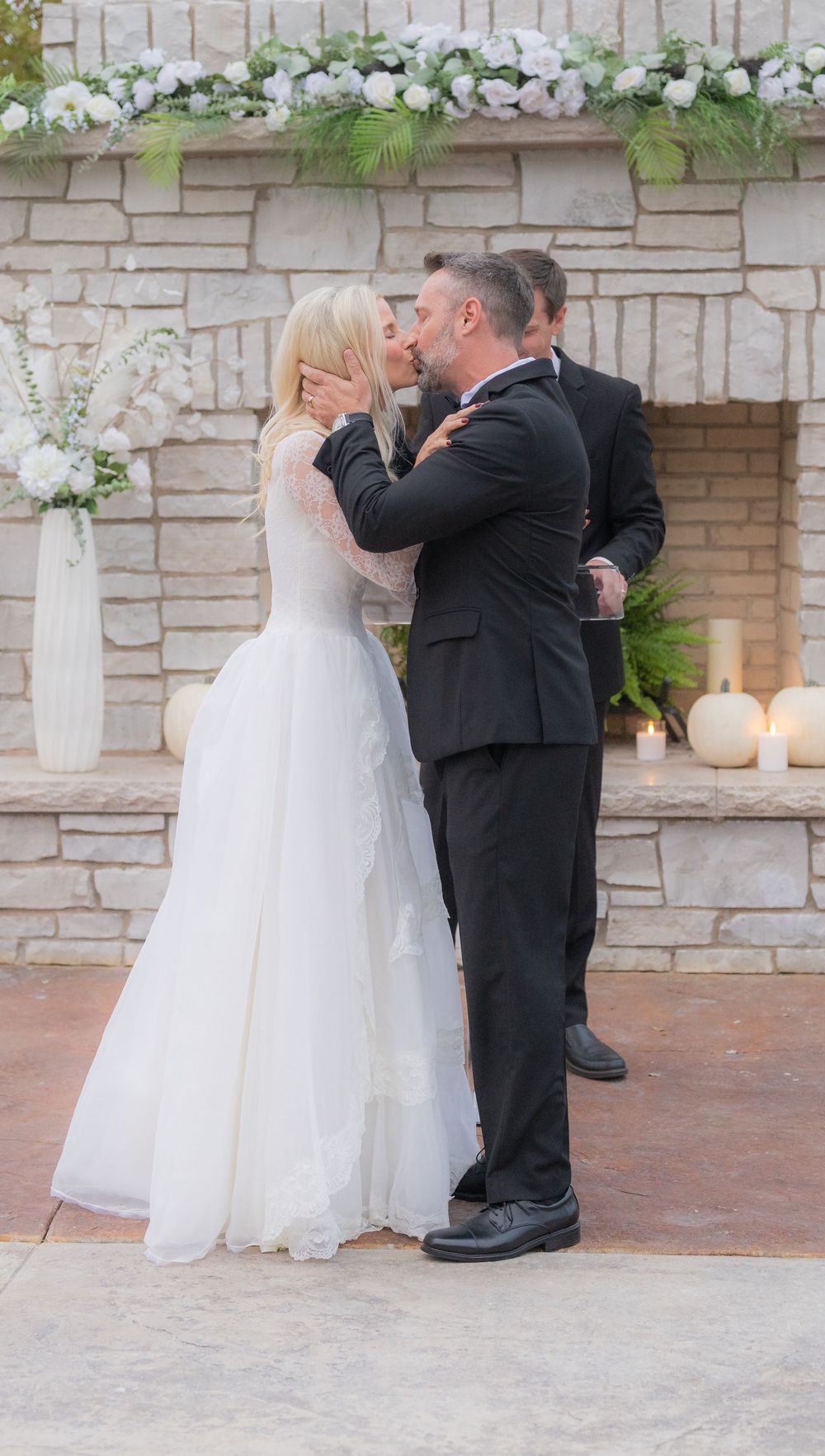 A couple in formal wedding attire shares a romantic moment during their outdoor ceremony at a stone-walled venue.