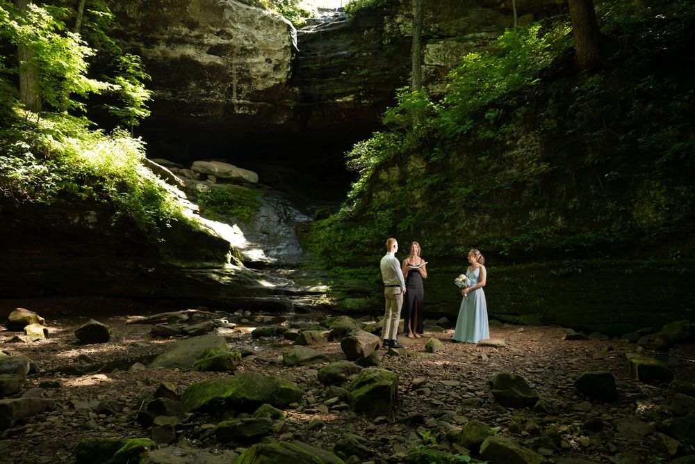 Couple exchanges vows with officiant in a lush rocky canyon with waterfall backdrop during an outdoor elopement ceremony.