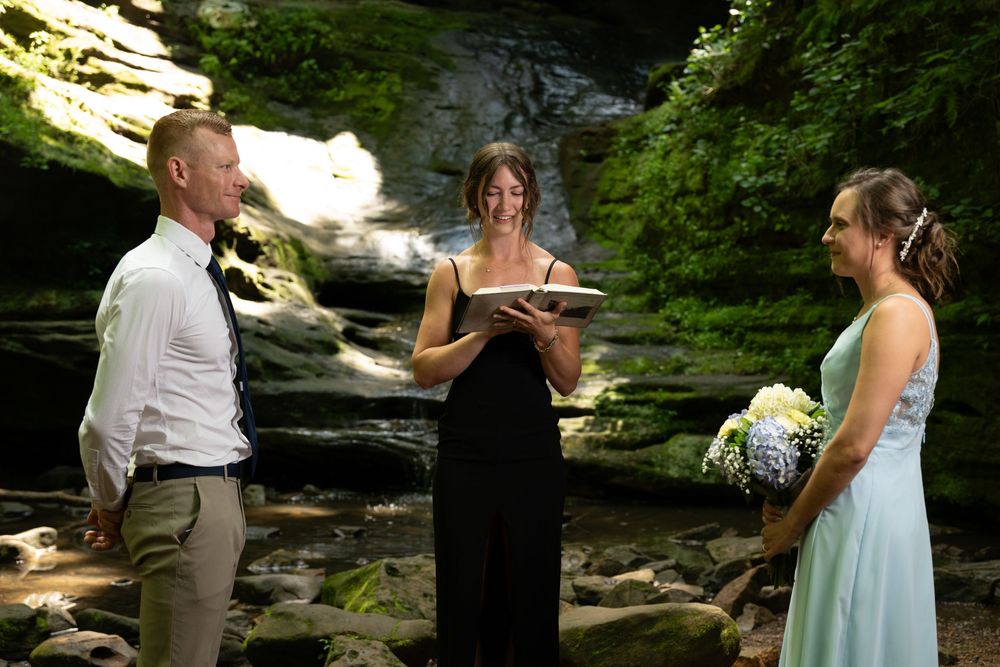 Outdoor wedding ceremony by a waterfall with officiant in black dress reading vows to couple in casual attire.