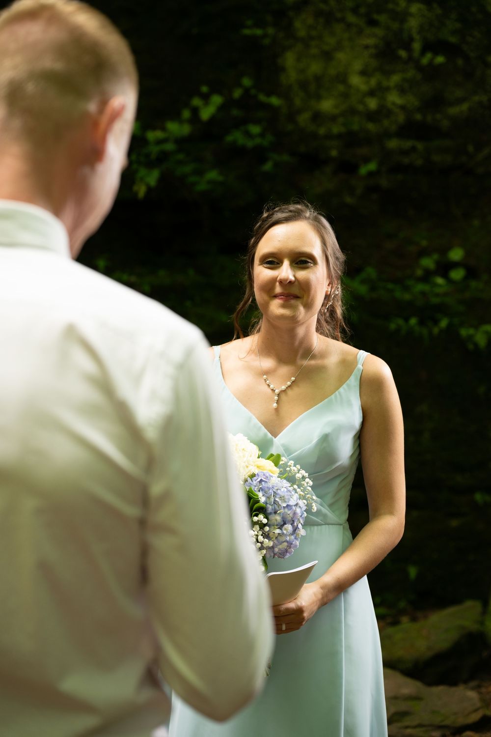 Bride in mint green dress holds bouquet, smiling at groom during outdoor wedding ceremony in lush forest setting.