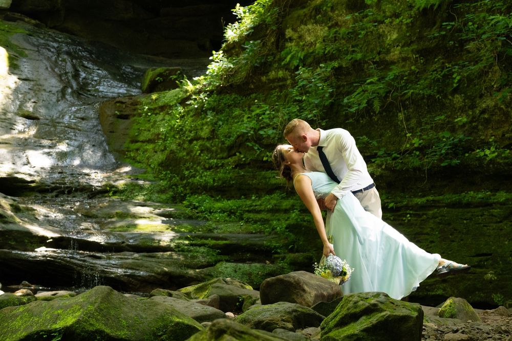 Bride and groom sharing a romantic dip kiss on mossy rocks near a waterfall in a lush green forest.
